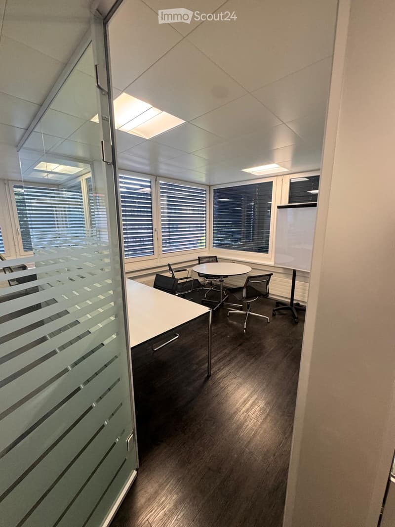 Office space with a round table, chairs, and a screen, featuring large windows with blinds and a glass partition.