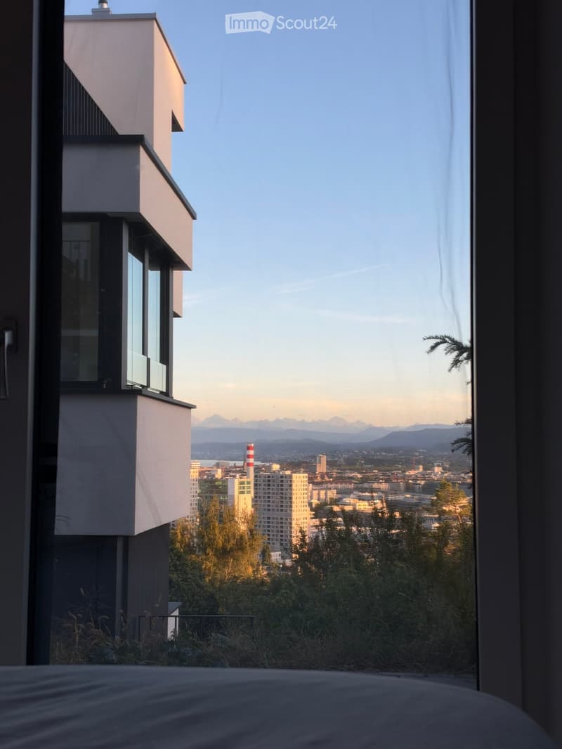 Bedroom with a large window offering a panoramic cityscape view, including buildings, trees, and distant mountains.