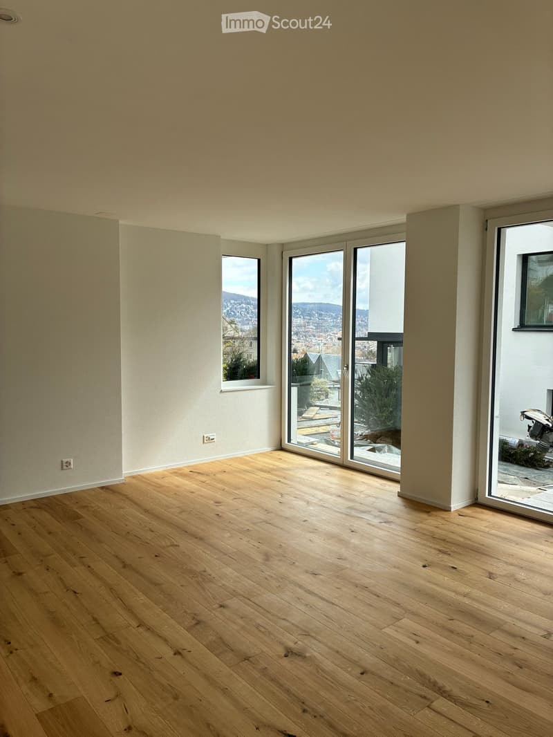 Empty room with light wooden flooring, white walls, and large windows offering a view of the city and surrounding landscape.