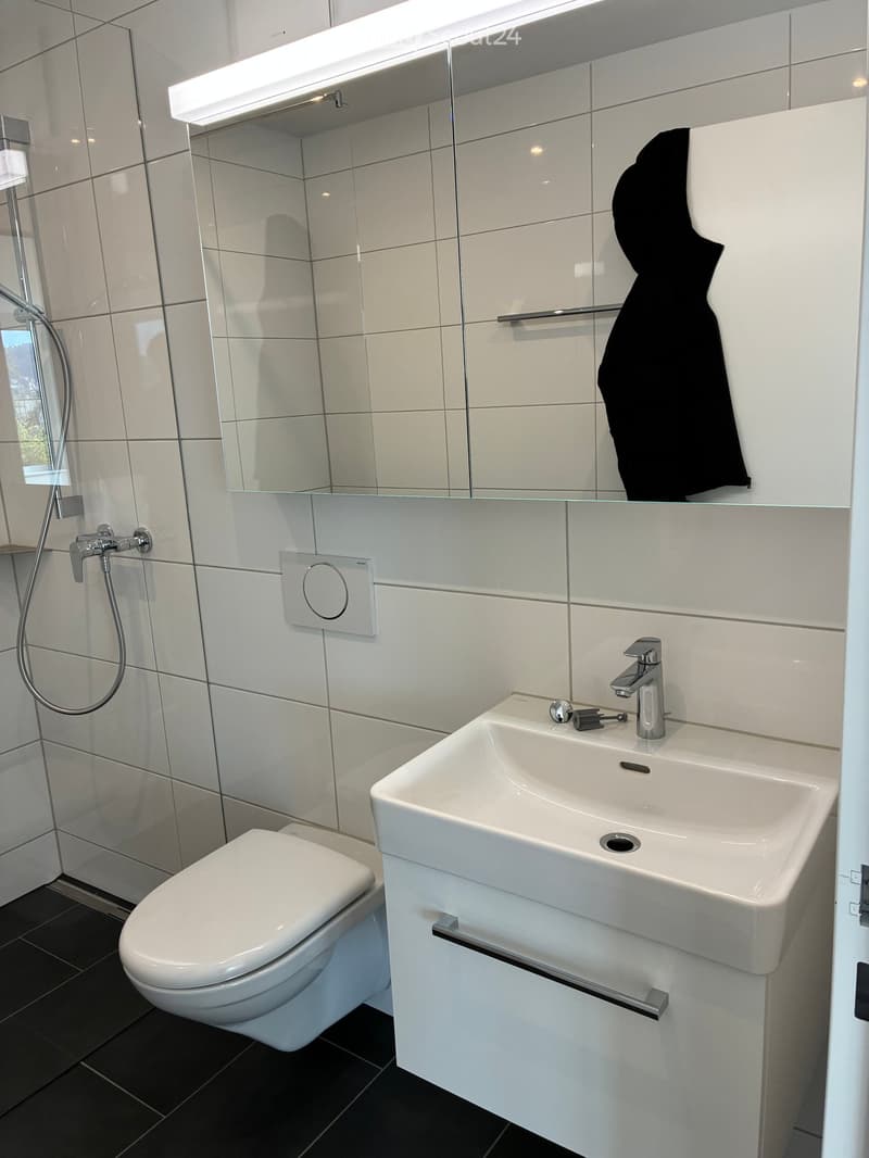 Modern bathroom with white tiles, a toilet, a sink with a modern faucet, a shower area, and a large mirror with built-in lighting. A black towel is hanging on a rack.