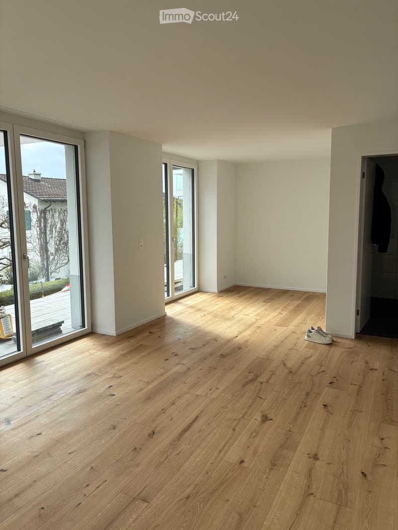 Empty room with light wooden flooring, white walls, large sliding glass doors leading to an outdoor area, and a pair of white sneakers placed near the entrance.