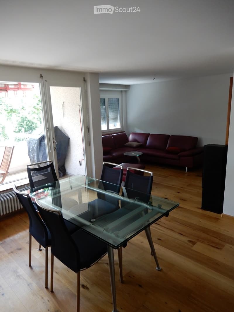 Dining area with a glass table and chairs, a red leather sofa, and a small round stool, connected to a living space with wooden flooring.