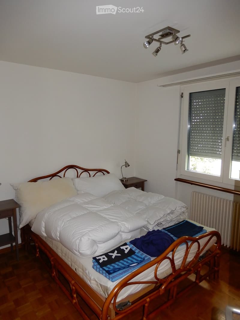 Cozy bedroom with a wooden bed, white bedding, nightstand, and a lamp. The room features wooden flooring and a radiator under the window.