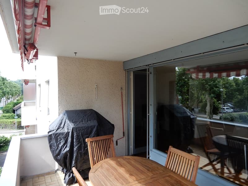 Balcony with wooden table and chairs, covered grill, sliding glass door leading inside, view of greenery and street.