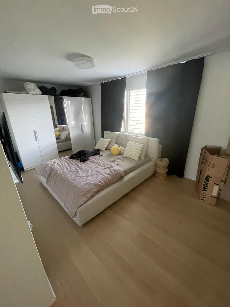 Modern bedroom with a large bed, white wardrobe, mirror, and a window with blinds. The room features light-colored wooden flooring and has some items like pillows, a ball, and a screen visible.