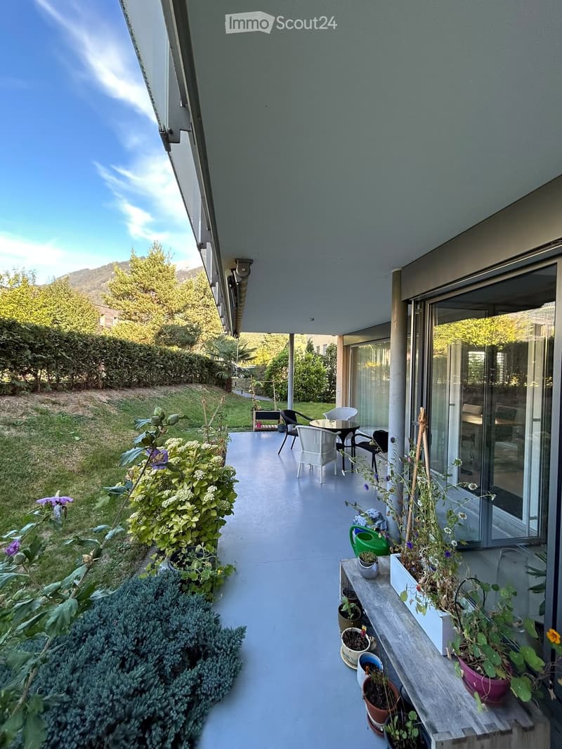 Balcony with a table, chairs, and several potted plants, overlooking a green garden area.