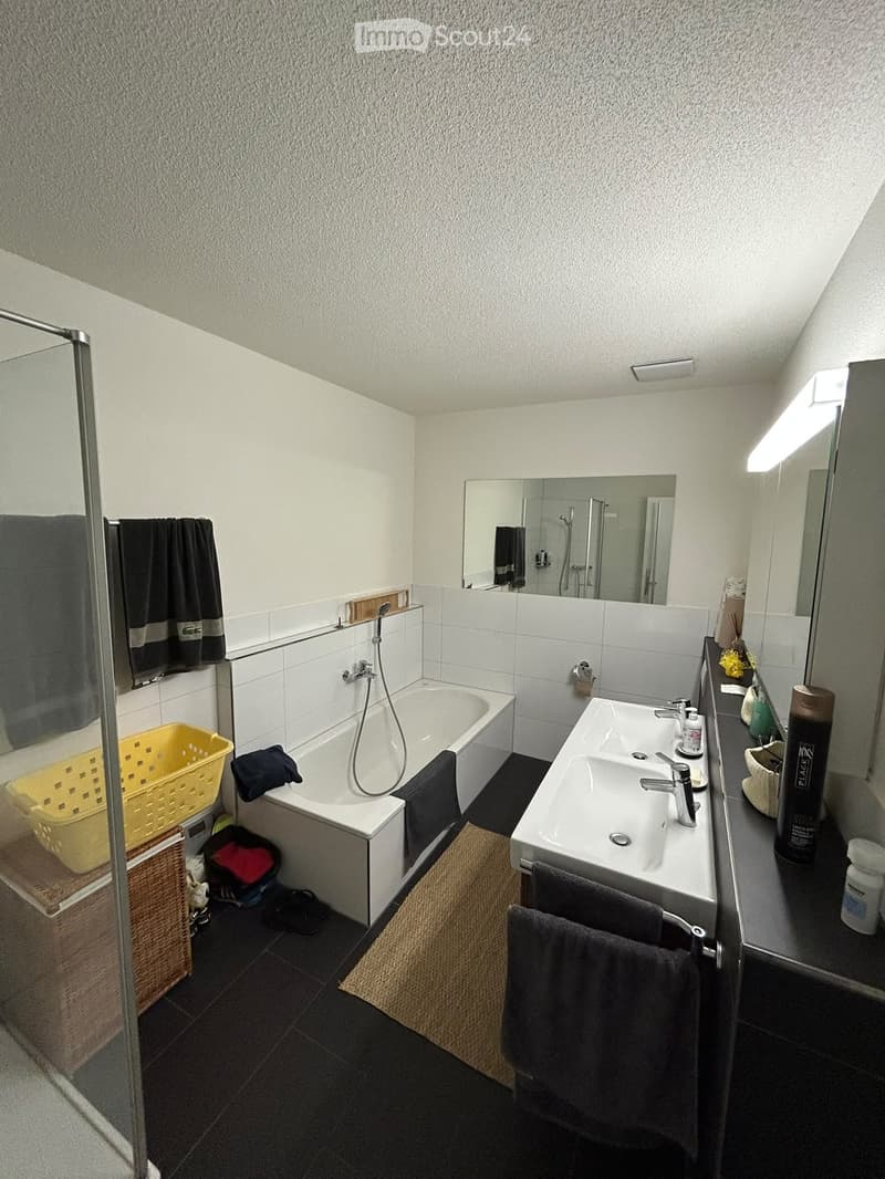Modern bathroom with a bathtub, double sink, large mirror, and towels. Includes a shower area with a glass enclosure and a yellow basket on the floor.