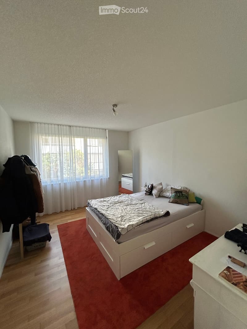 A well-lit bedroom with a white bed, nightstand, mirror, and a red rug on the floor. The room features wooden flooring and a large window with sheer curtains.