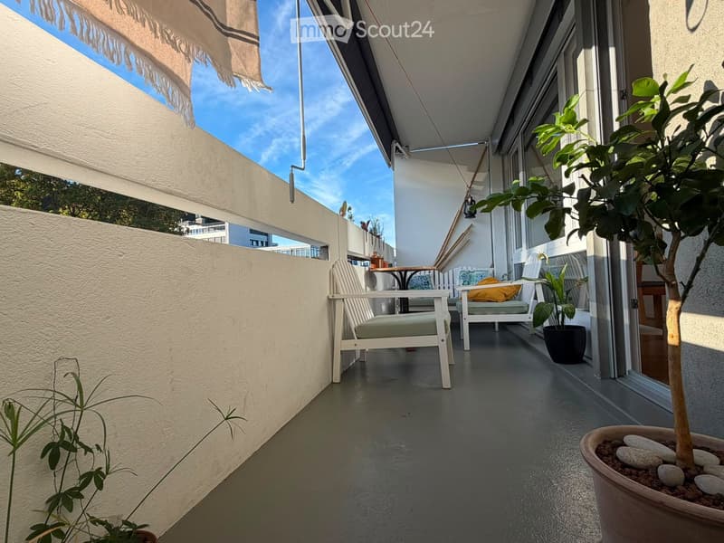 Balcony with grey floor, white chairs, small table, potted plants, and a large awning providing shade.