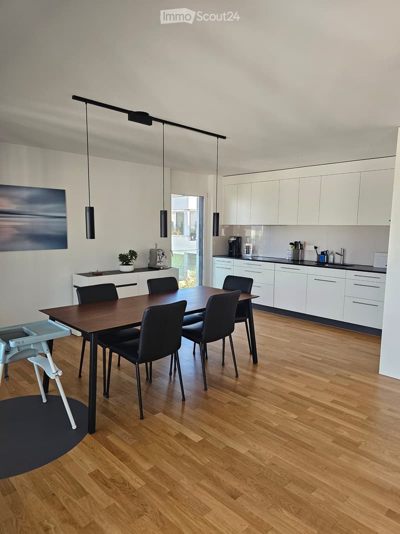 Modern kitchen and dining area with light wood flooring, white cabinets, a wooden dining table with black chairs, and a baby chair. Features include a coffee maker, potted plant, and pendant lights.
