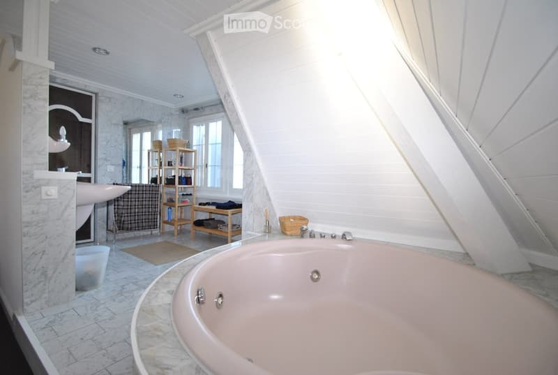 Bright bathroom with a large, freestanding bathtub, a sink with a mirror, and a wooden shelf holding various items. The room features white wooden paneling and large windows allowing natural light to fill the space.