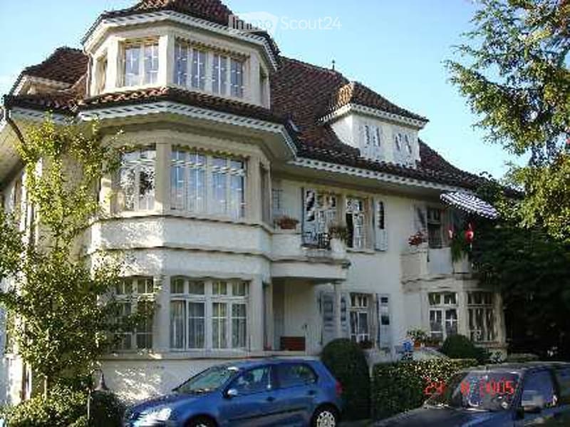Two-story house with white exterior, multiple windows, and a balcony on the upper floor. A blue car is parked in front of the house.