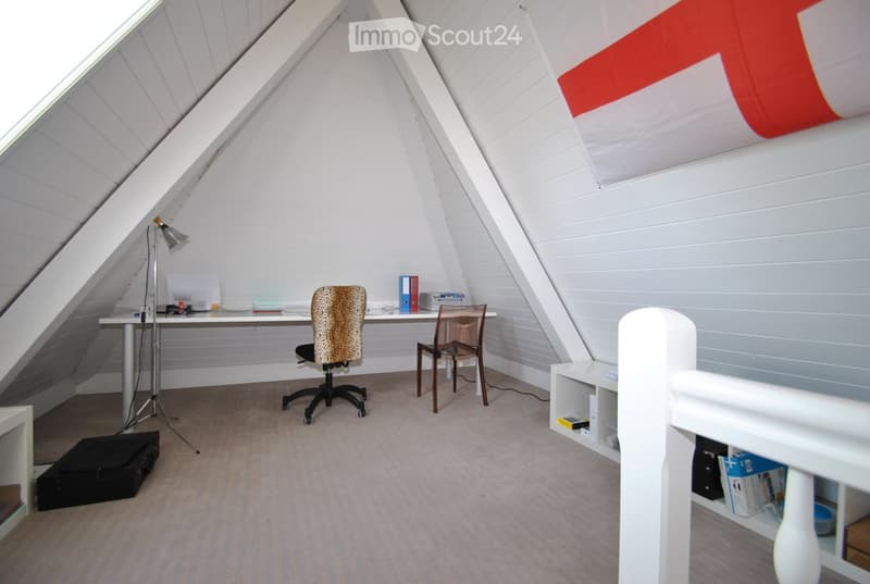 Attic room with slanted ceilings, white walls, and wooden trim. Features a desk with a chair, a floor lamp, and a small shelf with books and storage boxes.