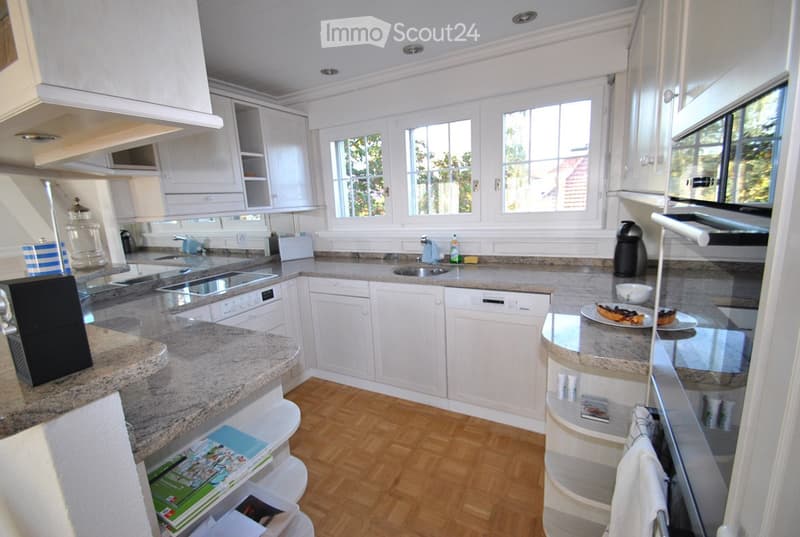 Modern kitchen with white cabinets, granite countertops, and wooden flooring. Features include a dishwasher, oven, sink, and various kitchen items on the counters.