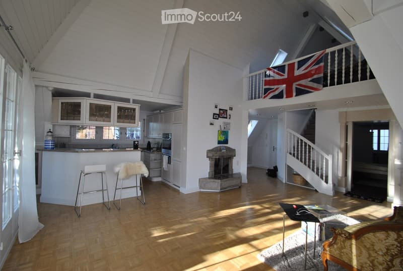 Bright, open-plan living and dining area with wooden flooring, white walls, and a fireplace. Features a sofa, coffee table, kitchen area with cabinets and appliances, and a staircase leading to an upper level.
