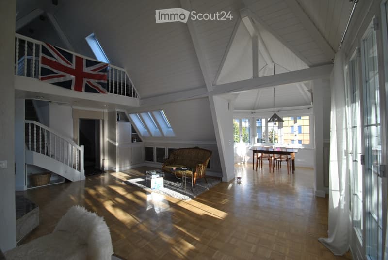 Bright, open-plan living and dining area with wooden floors, high ceilings, large windows, and skylights. Features a patterned sofa, coffee table, dining table with chairs, and a fluffy white rug.