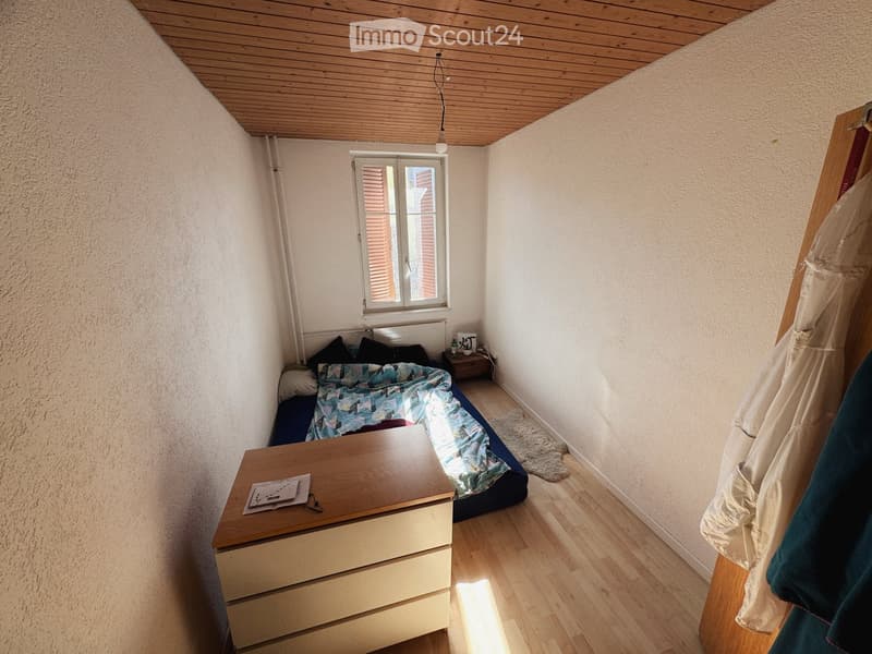 Cozy bedroom with wooden floor, slanted wooden ceiling, bed with colorful quilt, nightstand, small table, and radiator under window.