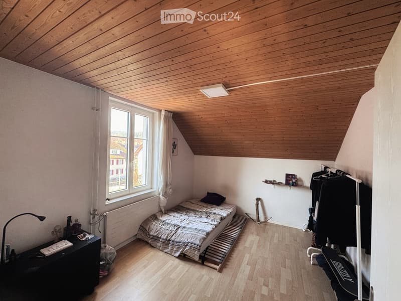 Cozy attic bedroom with wooden slanted ceiling, bed, nightstand, lamp, window with curtain, radiator, shelf, and clothing rack.
