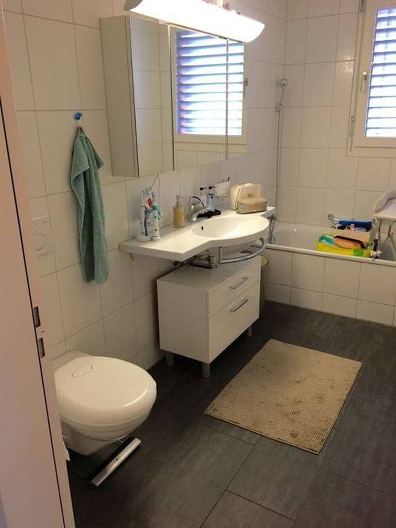 A modern bathroom with a white sink, mirror, cabinet, toilet, and bathtub. A towel hangs on the wall, and personal care items are placed on the sink counter. The floor is tiled, and a bathmat is placed in front of the bathtub.