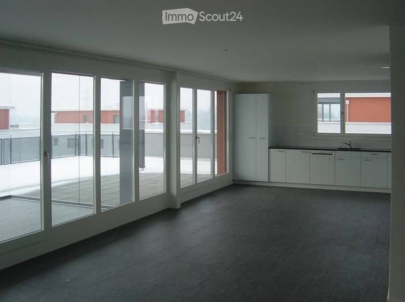 Empty room with large glass sliding doors leading to a balcony, white kitchen cabinets, and a sink visible in the background.