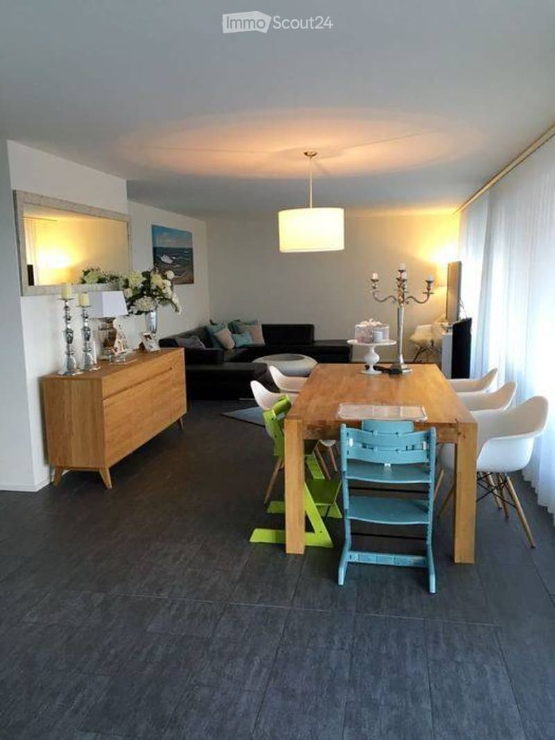 Modern living and dining area with a dark wood floor, a black sofa, a wooden dining table with chairs, a wooden cabinet, and decorative items including candles and a vase.