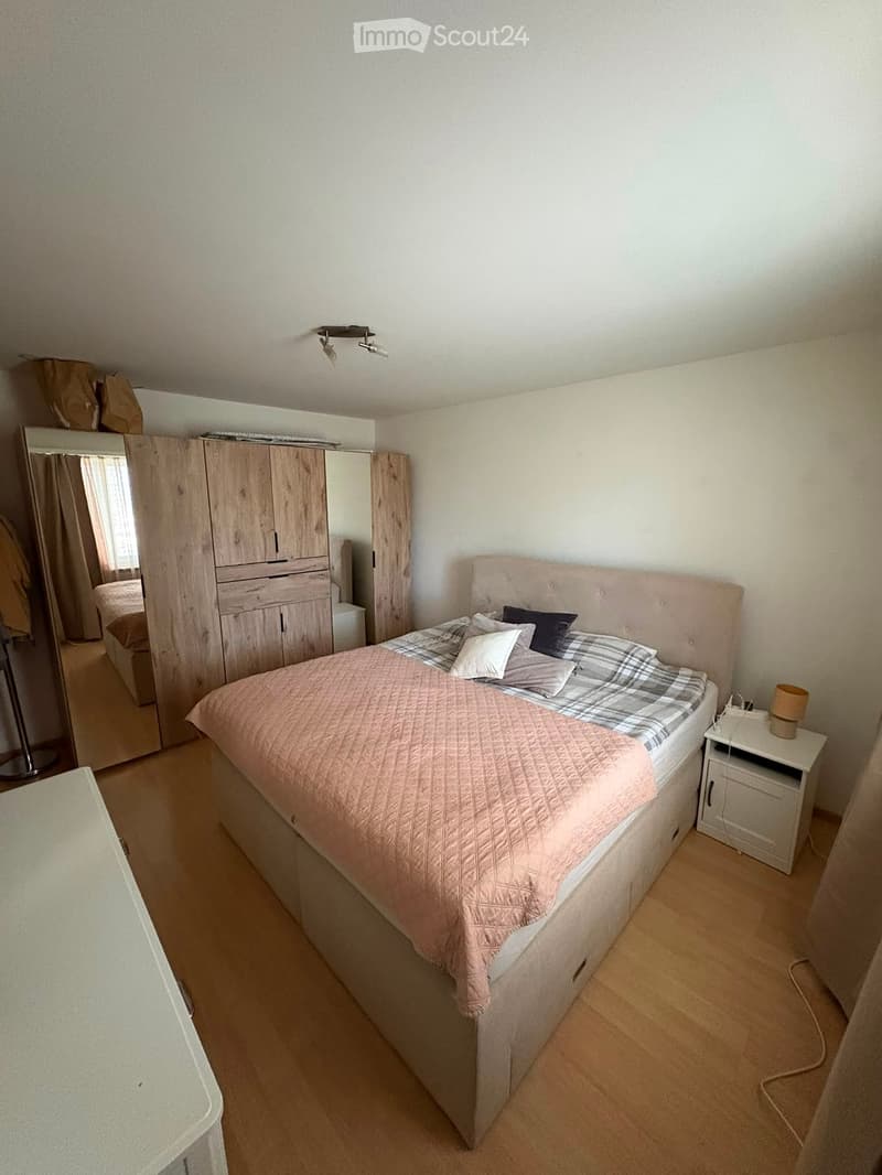 Cozy bedroom with a large bed, nightstand, lamp, wardrobe, and mirror. The bed is neatly made with a pink quilt and plaid blanket.