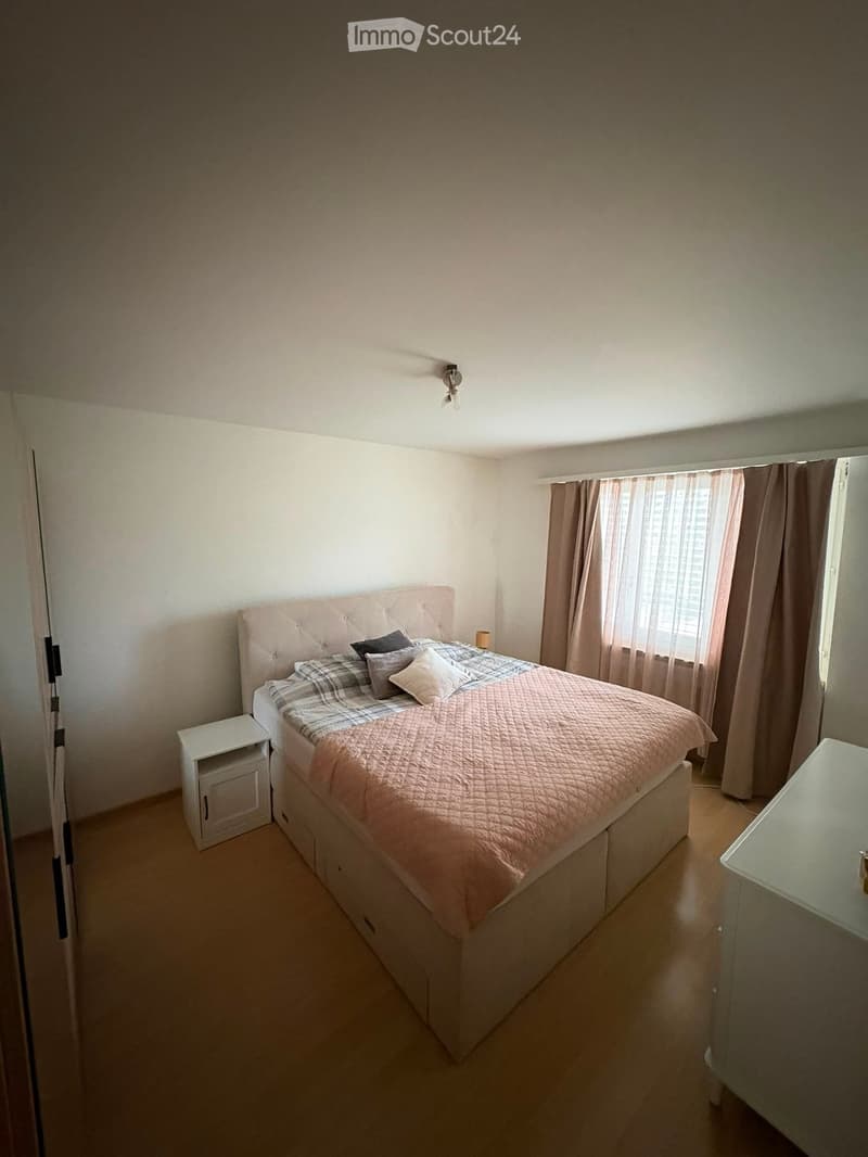 A well-lit bedroom with a large bed, nightstand, and a small table near the window with curtains.