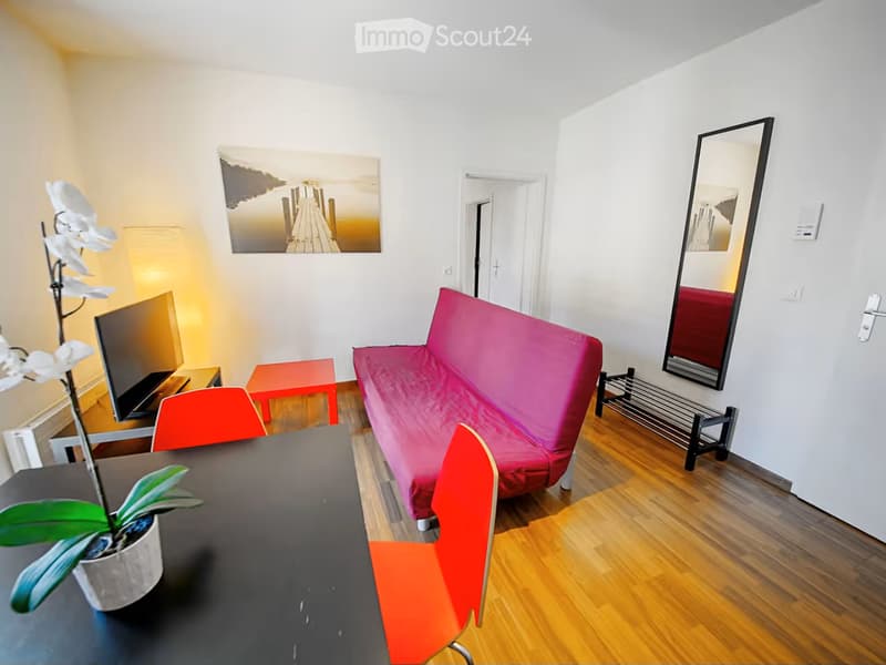 A modern, well-lit living room with a red sofa, a black coffee table, colorful chairs, a large mirror, and a TV screen. The room features wooden flooring and a potted plant on the table.