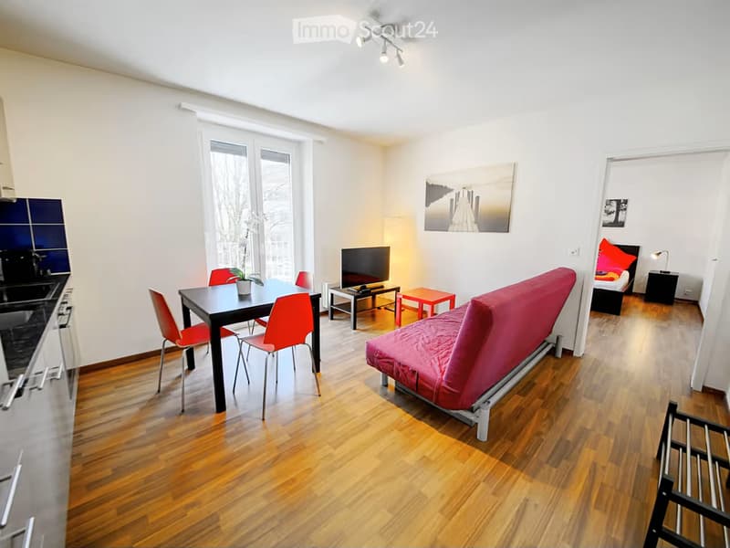 A modern, open-concept living and dining area with a red sofa, black dining table with red chairs, a television, and a small bedroom area with a bed and nightstand.