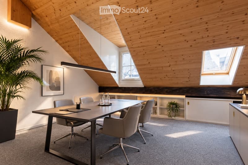 Modern office space with a large wooden table, several chairs, built-in cabinets, pendant lighting, and a large potted plant.