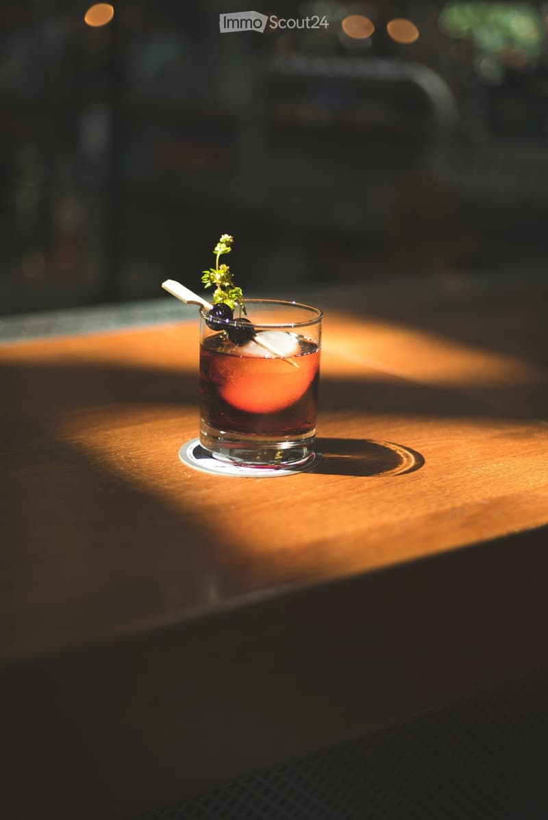 A cocktail in a short glass with ice, olives, and a herb garnish on a wooden surface.