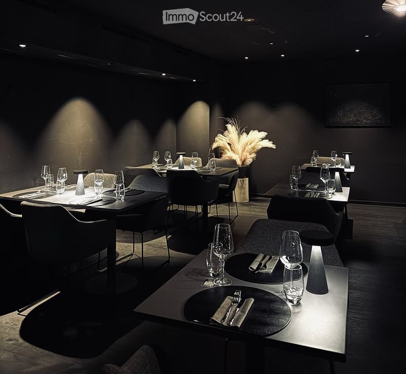 Elegant restaurant interior with dark walls, modern tables and chairs, wine glasses, plates, and cutlery set for dining.
