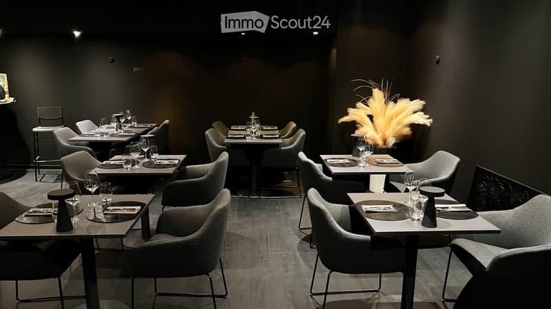 Modern restaurant interior with multiple tables and chairs, wine glasses, plates, and cutlery set for dining. A decorative vase with feathers is placed on one of the tables.