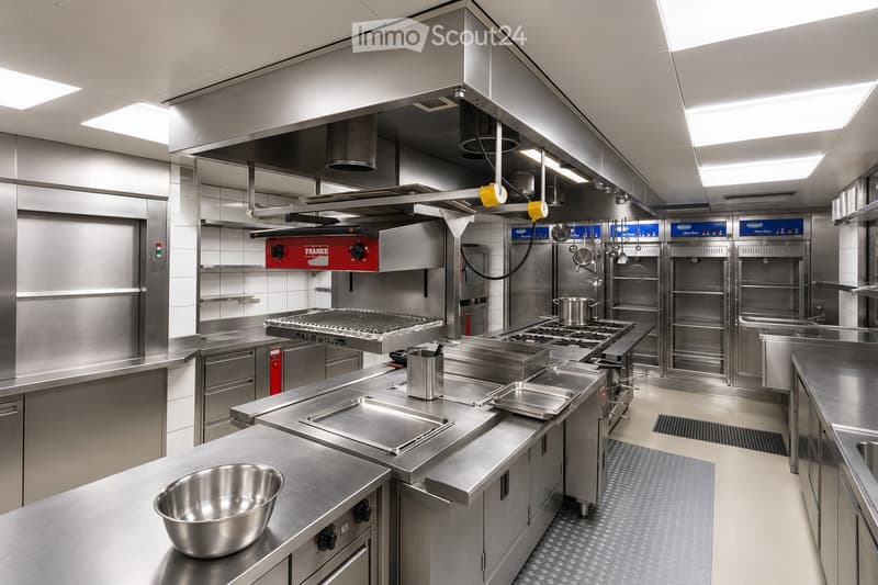 Commercial kitchen with stainless steel surfaces, multiple cooking stations, and professional-grade appliances.