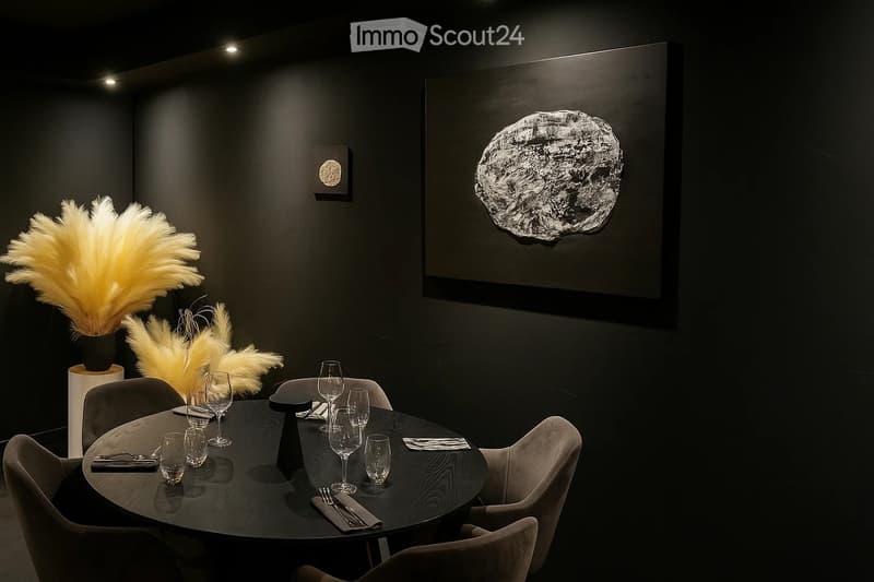 Elegant dining room with a round wooden table set for six, surrounded by modern chairs. The table is adorned with wine glasses, cutlery, and napkins. A large, sculptural fountain is mounted on the wall, and decorative feather arrangements in vases add a t