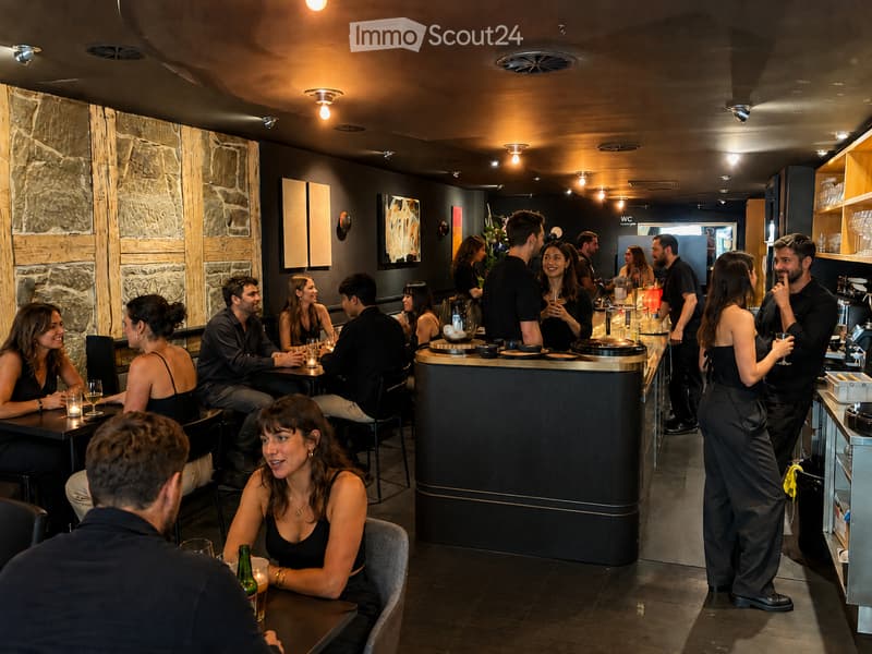 A modern restaurant interior with patrons seated at tables and standing near the bar, featuring dark wooden tables, chairs, and a central bar area with stools. The decor includes stone and wood paneling, modern lighting, and artwork on the walls.