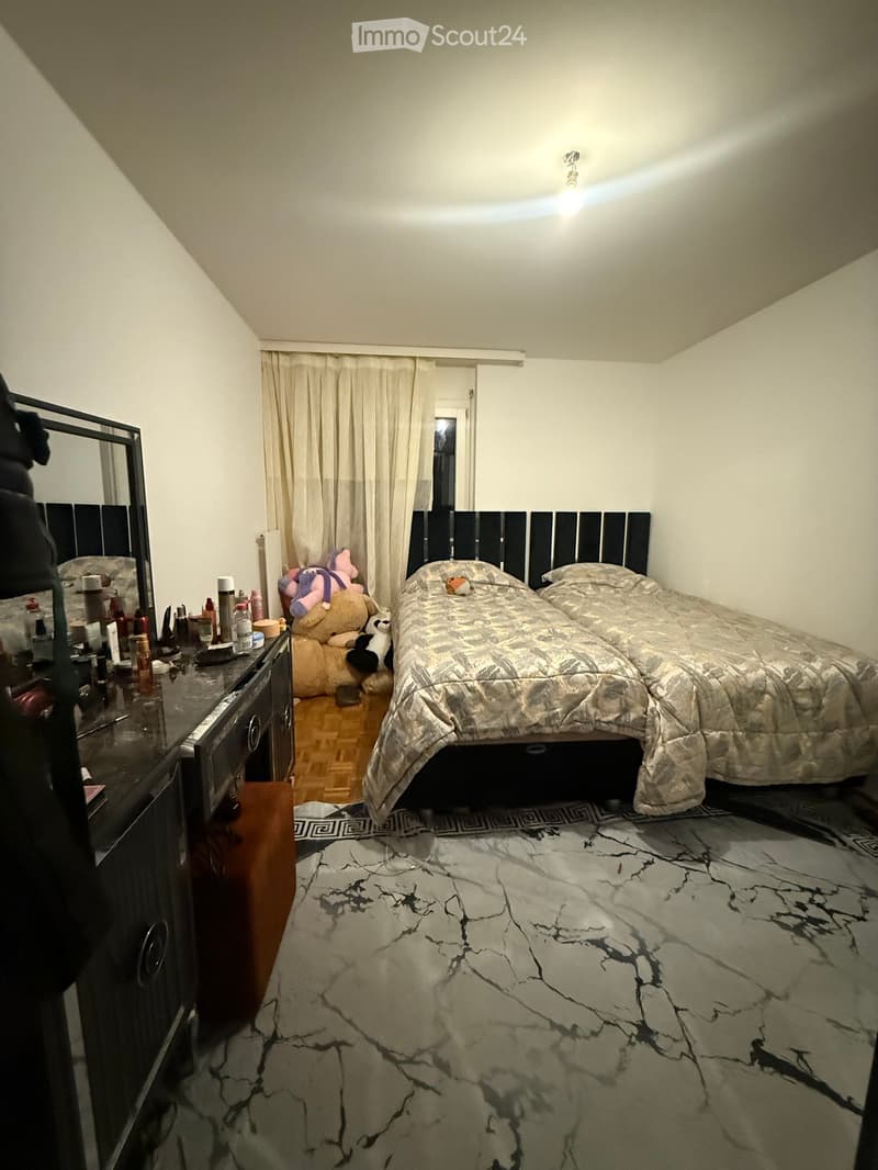 A cozy bedroom with a large bed, a vanity with various items, and a mirror. The room features a marble-patterned rug and a slanted ceiling with a light fixture.