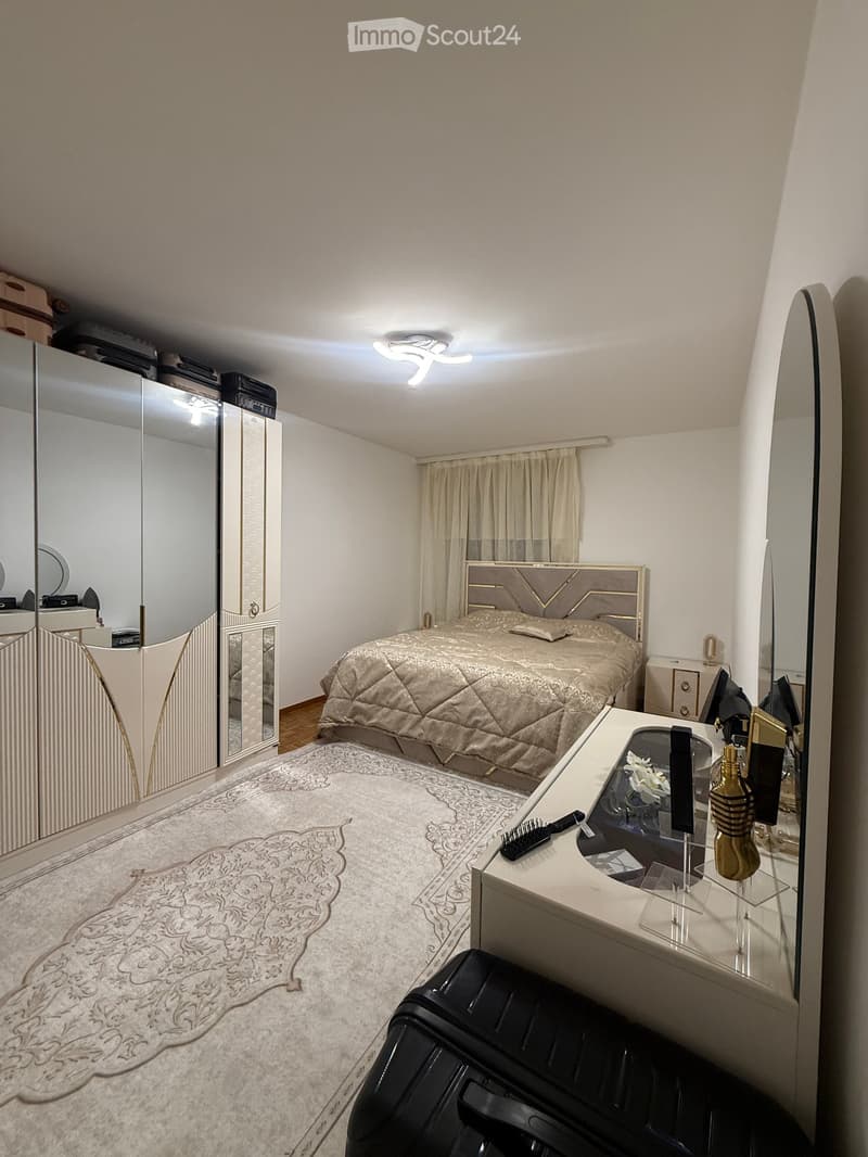 Modern bedroom with a large bed, beige bedding, a patterned rug, a vanity with perfume bottles, and a large mirror on the wall.