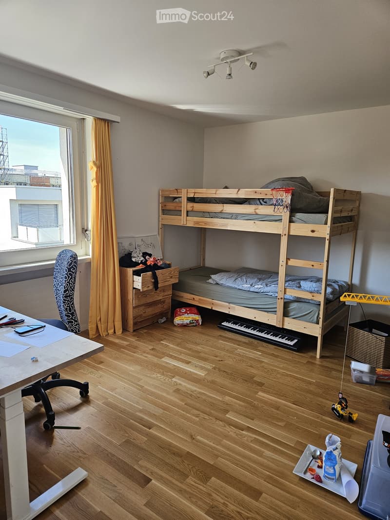A small, multi-functional room with a lofted bed, desk, and various items including a keyboard, toy car, and a mirror. The room features wooden flooring and a window with a view of a construction site.