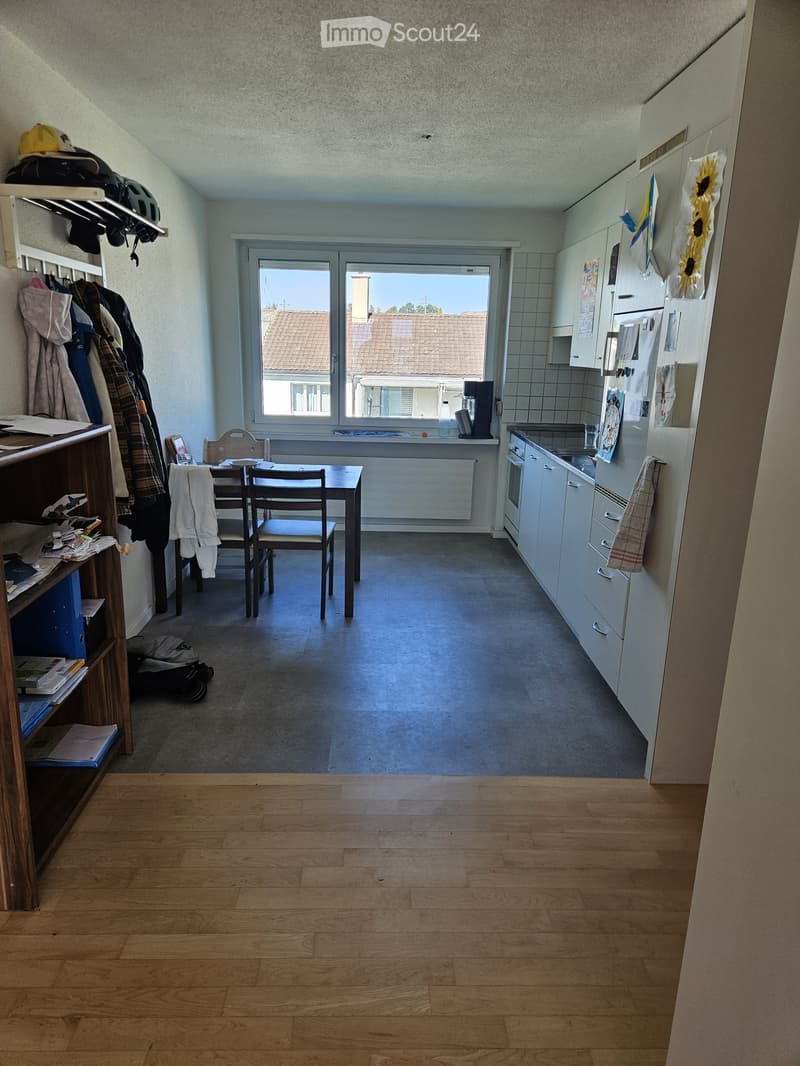 Empty room with wooden flooring, white walls, and a large window offering a view of the outside. The room contains a few storage shelves and a small table, but no furniture.