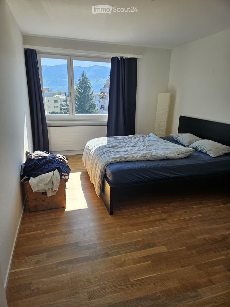 Bedroom with a large bed, nightstand, and clothing items on the floor. The room features wooden flooring and large windows with a scenic view.