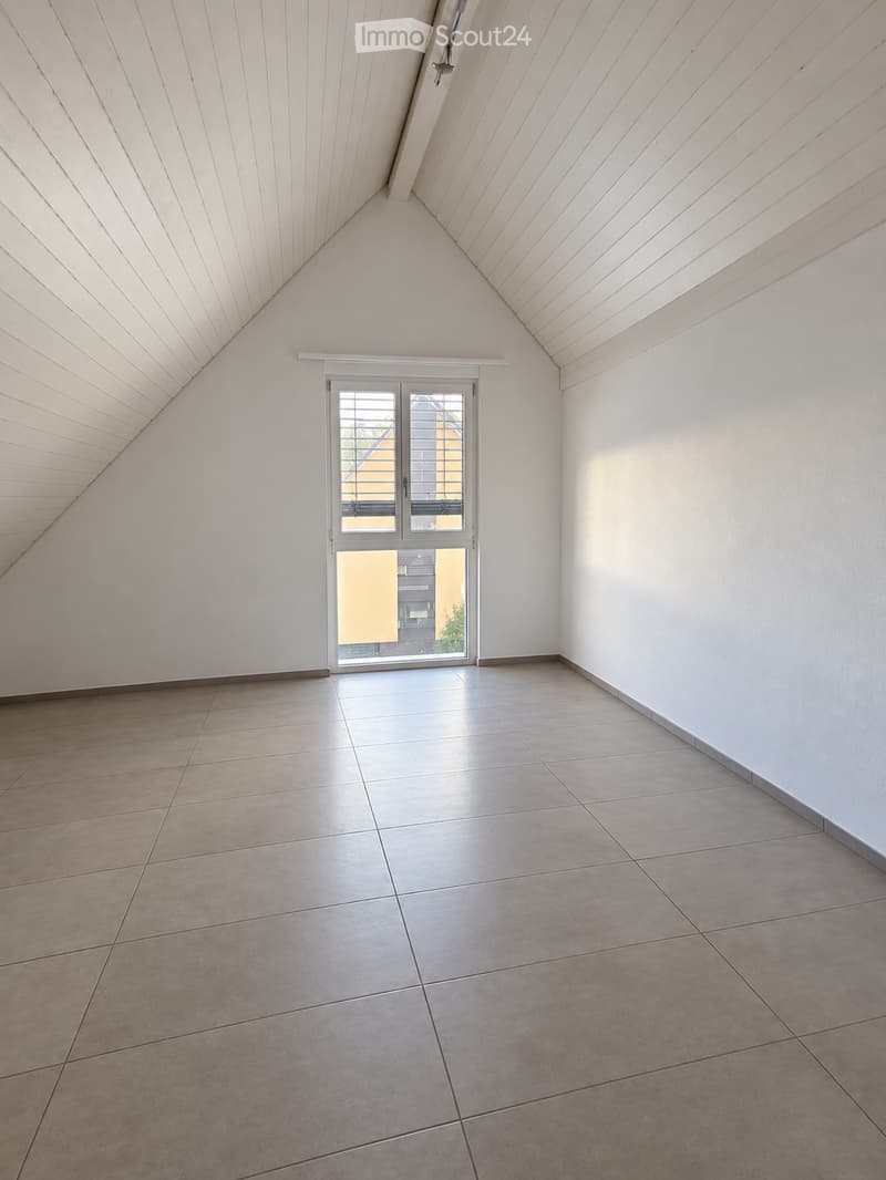 Empty room with white walls, tiled floor, and a large window with blinds. The ceiling has a unique slanted design.