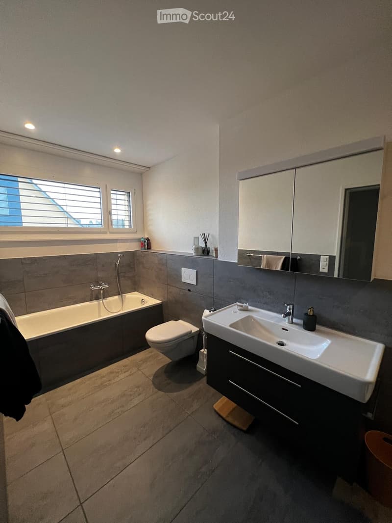 Modern bathroom with a bathtub, toilet, sink, and mirror. Features include a large window with blinds, grey tiled walls, and a dark wooden cabinet under the sink.