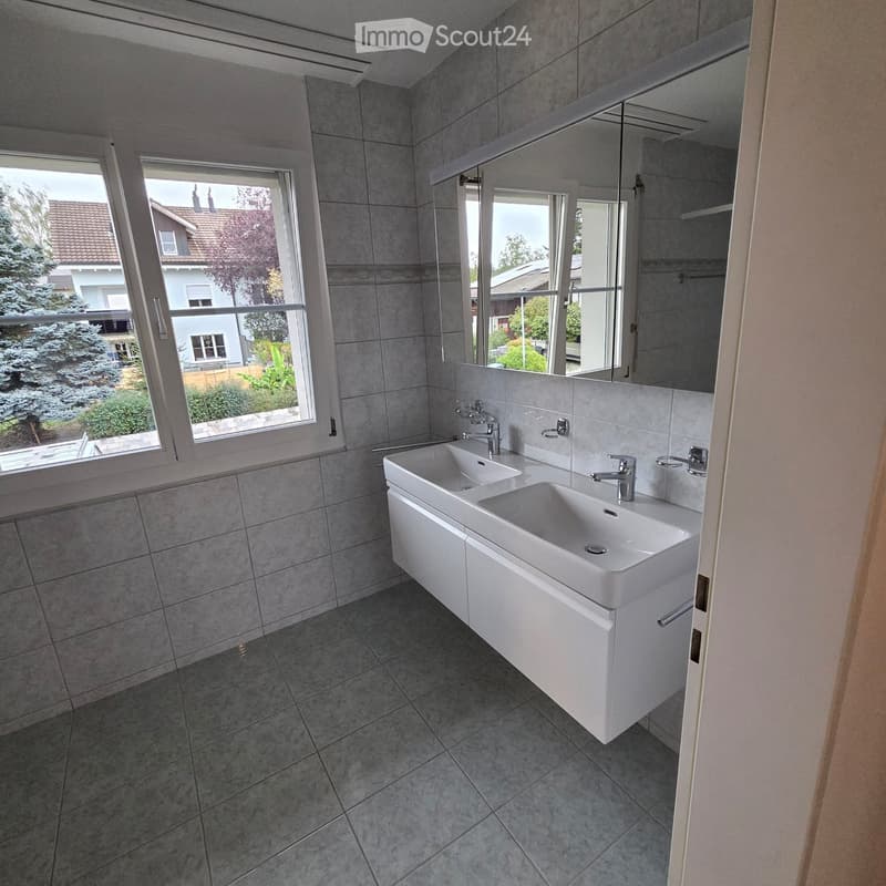 Modern bathroom with grey tiled walls and floor, double sink with modern faucets, large mirror, and large windows offering a view of the garden and neighboring house.