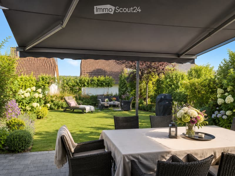 A well-maintained garden with a covered patio area, featuring a dining table with chairs, lounge chairs, a grill, and lush greenery.