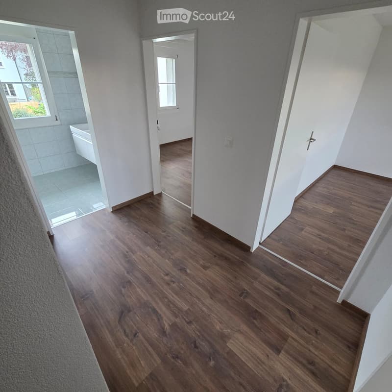 Empty hallway with wooden flooring, white walls, and doors leading to other rooms, including a bathroom with a bathtub visible through an open door.