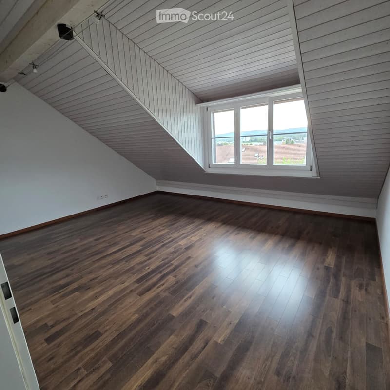 Empty attic room with wooden flooring, white paneled walls, and a large window offering a view of the surrounding area.