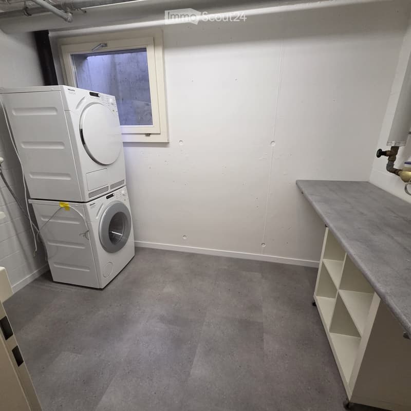 Small bathroom with a washing machine and dryer stacked, a shelf on the right side, and a window above the machines.