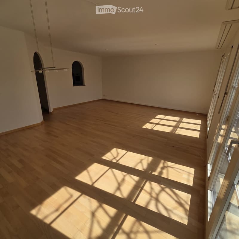 Empty room with light wooden flooring and white walls, featuring a large window with blinds and a small arched opening in the wall.