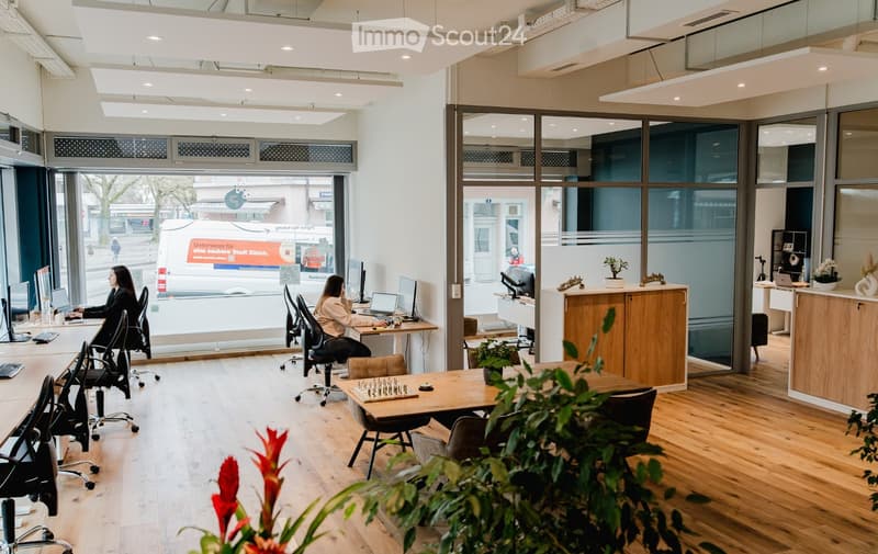 Spacious office space with large windows, wooden floors, ergonomic chairs, desks, monitor, and a game board on a table. Office furniture, potted plants, and equipment.